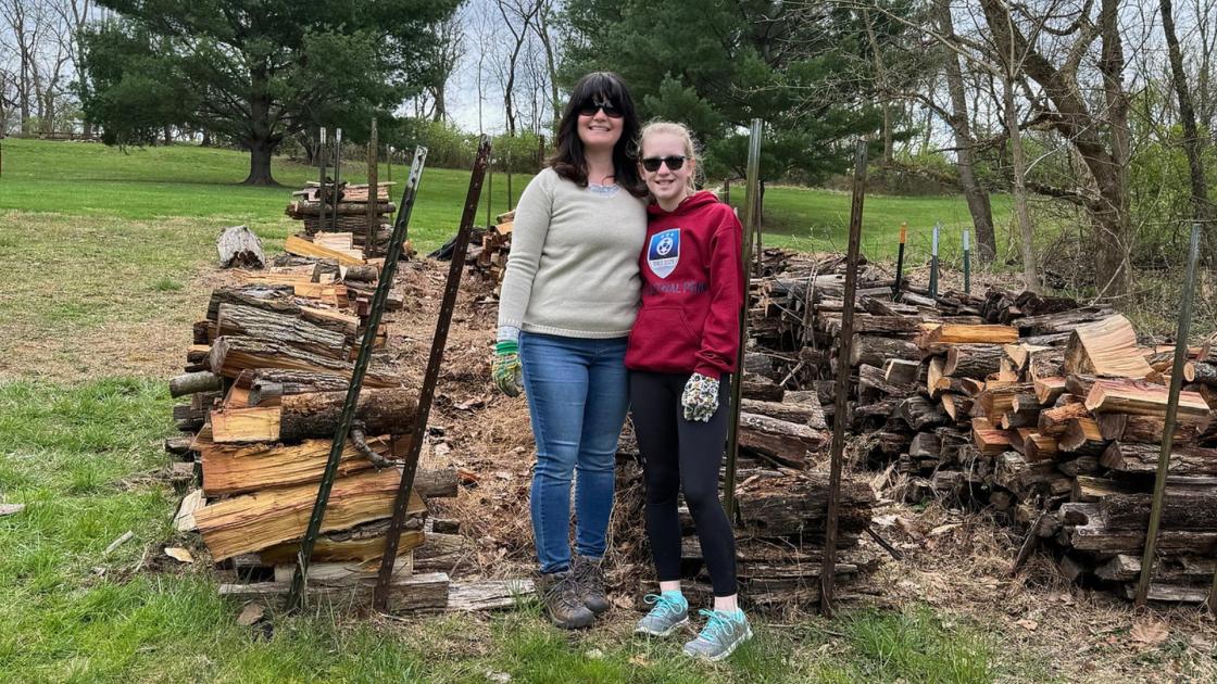 A woman and her daughter pose in front of a pile of wood that they helped stack